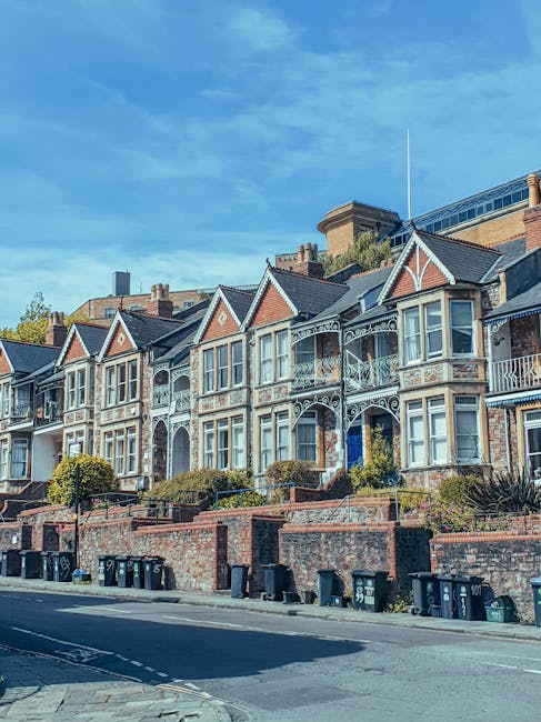 A row of traditional Victorian-style terraced houses with ornate white detailing and bay windows, situated on a slightly elevated street with a brick retaining wall and small garden beds in front. The